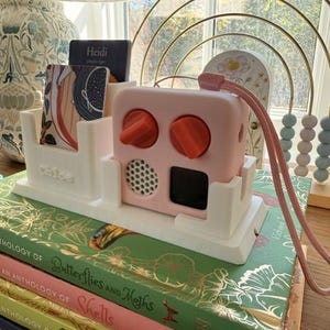 May include: A white plastic stand holds a pink audio player with two orange knobs, a circular speaker grille, and a black screen. The stand features a card slot containing a card labelled Heidi and a base embossed with the name Chloe.