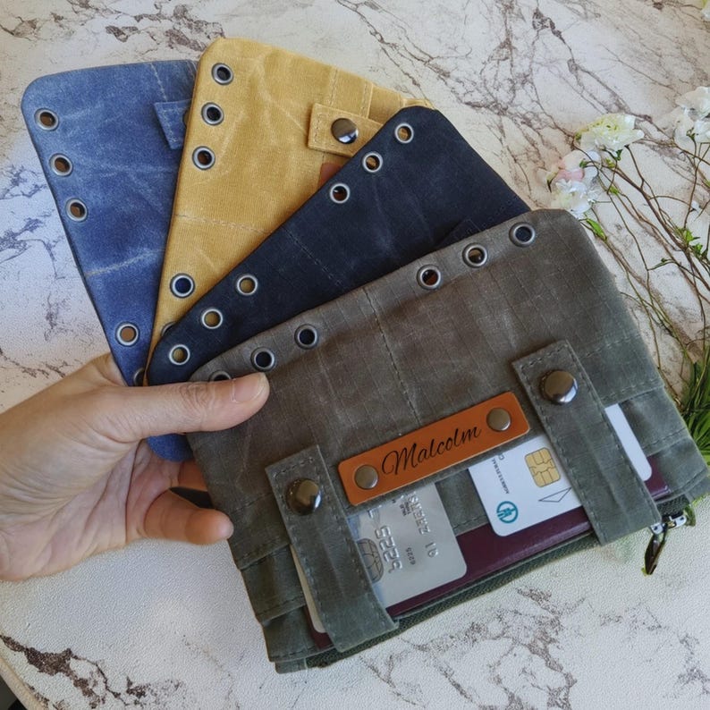 Op de afbeelding: Een verzameling canvas portemonnees in verschillende kleuren, waaronder blauw, mosterdgeel en olijfgroen. Elke portemonnee heeft metalen oogjes, drukknoopsluitingen en een leren label met de naam "Malcolm". De portemonnees hebben kaartsleuven en een ritsvak.