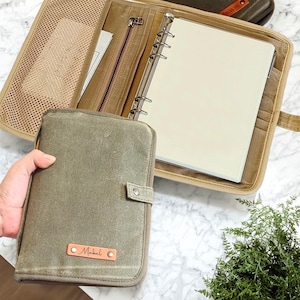 May include: An open olive green canvas binder with a zipper pocket and a mesh pocket. A closed matching canvas binder with the name "Mabel" on a leather tag is in the foreground. The binder is on a marble surface.