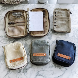 Könnte beinhalten: Eine Auswahl an Canvas-Reißverschlusstaschen und -Organizern in verschiedenen Farben. Ein offener Organizer zeigt einen Kalender für 2024 und einen Schlüsselhalter. Kleinere Taschen sind beige, grau und marineblau, jede mit einem Leder-Namensschild.