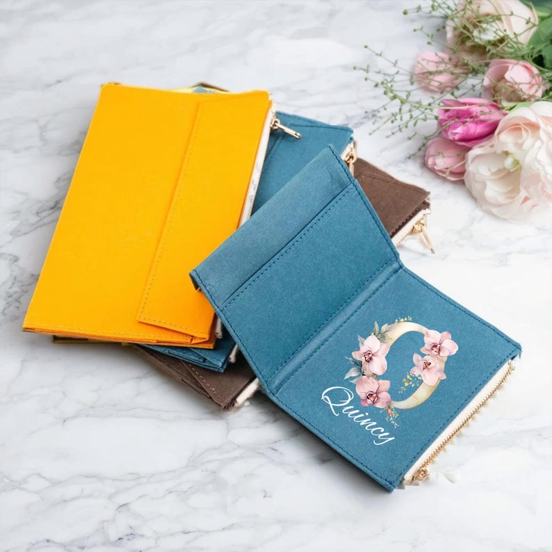 May include: A collection of colorful zippered wallets in shades of yellow, teal, and brown. One teal wallet is open, revealing a floral initial design with the name "Quincy" in script. The wallets are arranged on a marble surface.
