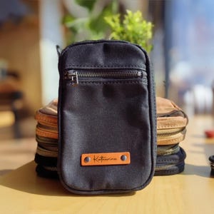 Könnte beinhalten: Schwarze Canvas-Tasche mit silbernem Reißverschluss und einem orangefarbenen Lederanhänger mit der Aufschrift "Katherine". Dahinter sind mehrere ähnliche Taschen in verschiedenen Farben gestapelt. Die Taschen haben eine rechteckige Form und einen Reißverschluss.