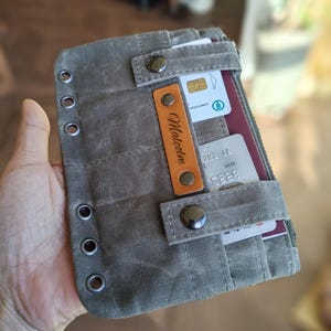 Op de afbeelding: Een grijze canvas portemonnee met meerdere kaartsleuven en een vak met ritssluiting. De portemonnee heeft een leren label met de naam "Malcolm" en metalen drukkers. Ontworpen om kaarten en andere benodigdheden in op te bergen.