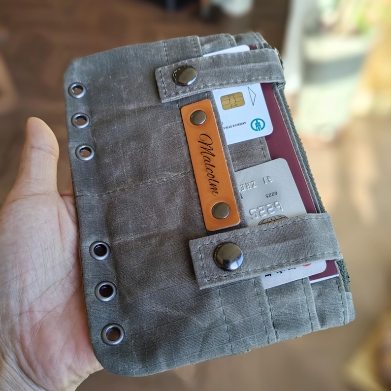 Op de afbeelding: Een grijze canvas portemonnee met meerdere kaartsleuven en een vak met ritssluiting. De portemonnee heeft een leren label met de naam "Malcolm" en metalen drukkers. Ontworpen om kaarten en andere benodigdheden in op te bergen.