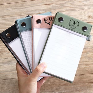 May include: A selection of personalised notepads with leather covers in black, teal, pink, and green. Each notepad features a different design, including lined and grid paper, and a monogram or graphic. The notepad covers are secured with silver snap closures.