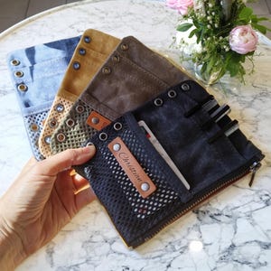 May include: A collection of canvas wallets in various colors, including blue, tan, and black. Each wallet features a mesh pocket, metal grommets, and a leather tag with the name "Christian". The wallets are open, revealing the interior and contents.