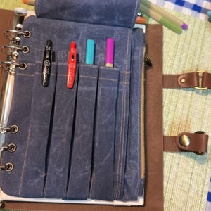 以下が含まれることがあります： 複数のポケットが付いた青いキャンバスのペンホルダー。黒いペン、赤いペン、2色のマーカーが入っています。ペンホルダーは、ジッパー開閉と革ストラップが付いた茶色の革張りのノートの中にあります。