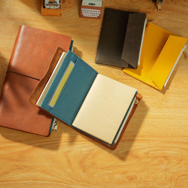May include: A collection of leather journals and pouches in various colors. The open journal reveals blank pages and a teal interior. A brown journal is open, displaying a teal interior with a clear card holder. Other pouches are black and yellow.
