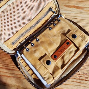Op de afbeelding: Open, met rits afgesloten, beige canvas organizer met een notitieboek en pen. De organizer heeft een mesh-vak, metalen ringen en een leren label met "Haley". Het notitieboek is vastgezet met drukknoppen.