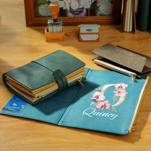 May include: A teal leather journal with a strap closure and a zippered pocket. The open journal reveals a teal cover with the name "Quincy" and a floral design. Other stationery items are in the background.