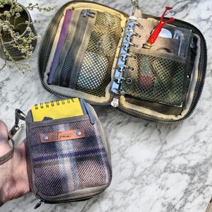 以下が含まれることがあります： 複数のコンパートメントを備えた、開いたオリーブグリーンのジッパー付きオーガナイザー。小さなリングバインダーとメッシュポケットが含まれています。「Irma」という名前の小さなお揃いのチェック柄ポーチと、黄色のメモ帳も表示されています。