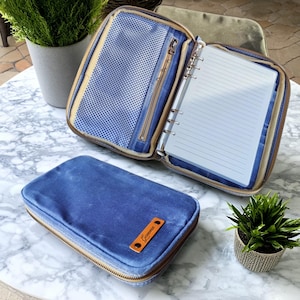 May include: A blue zippered binder case with a matching notebook. The case is open, revealing a lined notebook and a mesh pocket. A smaller, closed case with a leather tag that reads "Gemma" is in front. The cases are on a marble table.