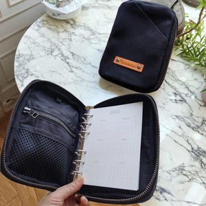 May include: Black zippered organizer with a 2025 calendar. The organizer has multiple compartments, including a zippered pocket and a mesh pocket. A smaller black organizer with a leather tag that says "Hermosa" is also visible.
