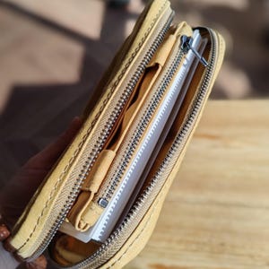 Op de afbeelding: Een lichtgele canvas etui of portemonnee, gedeeltelijk open om een notitieboek en binnenvakken te onthullen. De etui heeft een metalen ritssluiting en wordt door een hand opengehouden.