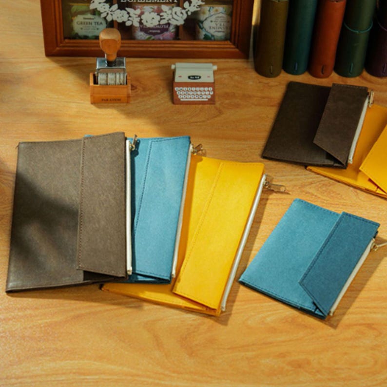 May include: A collection of colorful zippered pouches in brown, blue, and yellow, arranged on a wooden surface. The pouches are rectangular with a simple design and a metal zipper. Other stationery items are in the background.