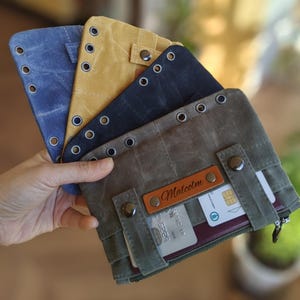Op de afbeelding: Een verzameling canvas portemonnees in verschillende kleuren, waaronder blauw, geel en olijfgroen. Elke portemonnee heeft metalen oogjes, drukknoopsluitingen en een leren label met de naam "Malcolm". De portemonnees zijn ontworpen om kaarten en andere benodigdheden in op te bergen.
