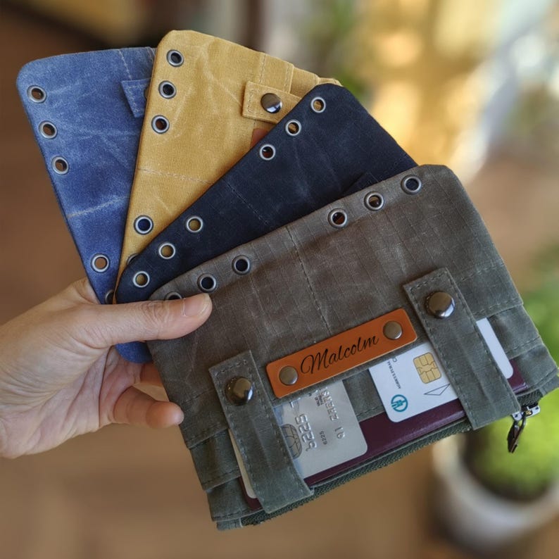 Op de afbeelding: Een verzameling canvas portemonnees in verschillende kleuren, waaronder blauw, geel en olijfgroen. Elke portemonnee heeft metalen oogjes, drukknoopsluitingen en een leren label met de naam "Malcolm". De portemonnees zijn ontworpen om kaarten en andere benodigdheden in op te bergen.