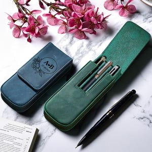 以下が含まれることがあります： 2つの革製ペンケース。1つはティール、もう1つはグリーンで、中にペンが入っています。ティールケースには花柄のデザインと日付入りのイニシャルがあります。黒いペンがグリーンのケースの隣にあります。背景にはピンクの花があります。