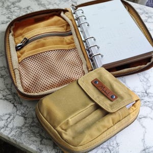 Könnte beinhalten: Geöffnete, hellbraune Organizer-Tasche mit mehreren Fächern, darunter eine Netztasche und ein Notizbuch. Eine kleinere, passende Tasche mit einem Lederetikett mit der Aufschrift "Michael" liegt davor.