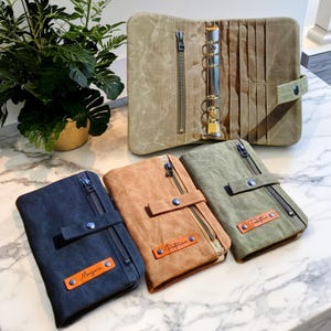 Peut inclure: Plusieurs classeurs en toile de différentes couleurs : bleu marine, marron et vert olive. Chaque classeur est doté d'une fermeture éclair, d'un bouton-pression et d'une petite étiquette en cuir avec un nom. Le classeur ouvert révèle un mécanisme à anneaux.