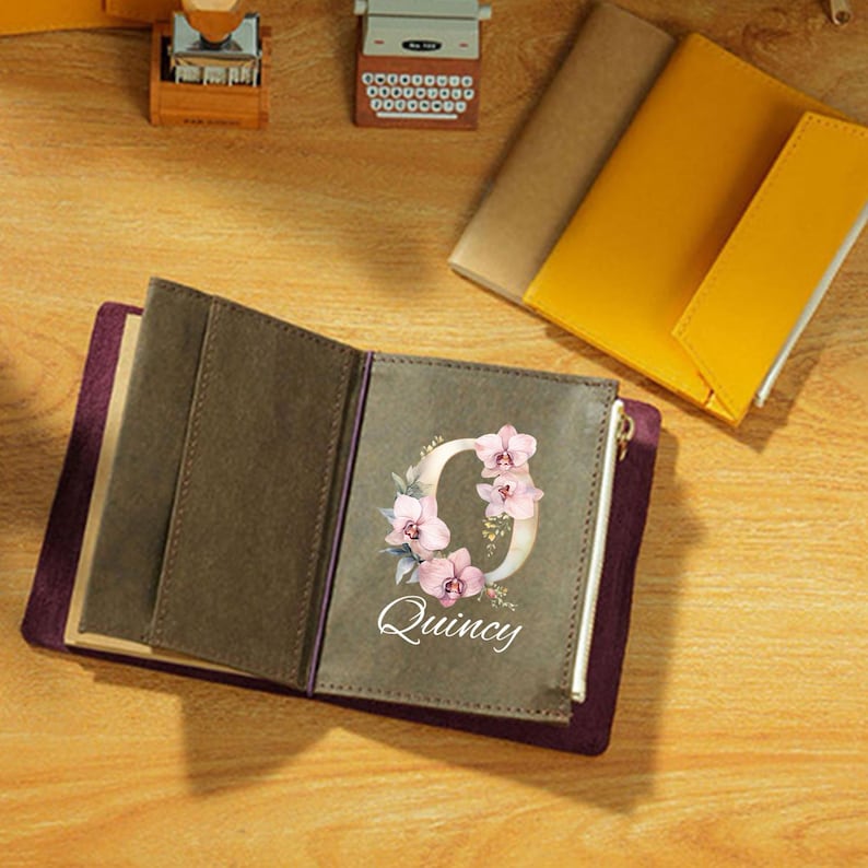 May include: Open journal with a personalized floral design. The cover is a deep purple with a gray interior. The journal features the letter "Q" adorned with pink orchids and the name "Quincy" in white script. Other journals and accessories are in the background.