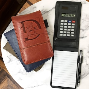 以下が含まれることがあります： 電卓とペンが付いた革製ノート。ノートは茶色で、「Damla」という名前が刻印されています。黒い電卓がノートに付いています。黒いペンはホルダーに入っています。ノートは白い大理石の表面にあります。