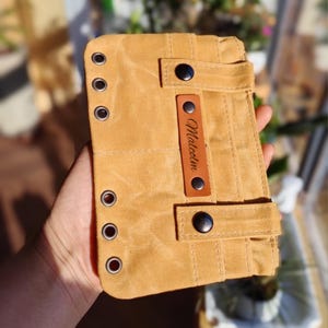 Op de afbeelding: Een bruin canvas portemonnee met een leren label met de tekst "Malcolm". De portemonnee heeft zwarte knoopsluitingen en metalen ogen. De portemonnee is opgevouwen en heeft een riemsluiting.