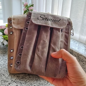 以下が含まれることがあります： フラップ開閉と金属アイレットが付いた、2つの茶色の生地の財布。前面の財布には「Samuel」という名前が印刷されています。財布は手で持たれています。