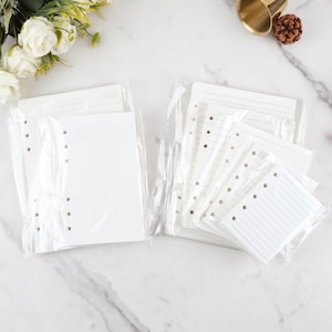Peut inclure: Deux piles de recharges de papier blanc pour un agenda, emballées dans du plastique transparent. Une pile est vierge, l'autre a des pages lignées. Chaque pile a des trous pré-perforés pour la reliure. Fleurs blanches et surface en marbre en arrière-plan.