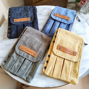 以下が含まれることがあります： 青、グレー、黄色の4色のキャンバス製ポーチ。それぞれに革製のネームタグが付いています。ポーチはプリーツのデザインで、側面に金属製のアイレットが付いています。各ポーチには、革製のタグに異なる名前が記載されています。