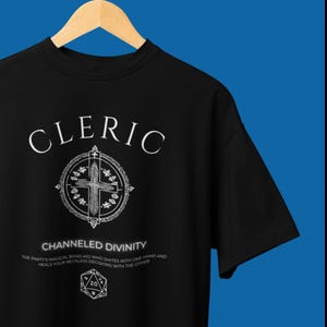 May include: Black t-shirt with a white graphic of a cleric symbol and the text "CLERIC" and "CHANNELED DIVINITY" with the text "THE PARTY'S MAGICAL BAND-AID WHO SMITES WITH ONE HAND AND HEALS YOUR RECKLESS DECISIONS WITH THE OTHER".