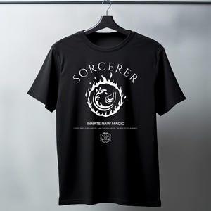 May include: Black t-shirt with a white graphic of a sorcerer's symbol. The symbol is a circle with flames and a wave inside. The text "SORCERER" is above the symbol and "INNATE RAW MAGIC" is below. The text "I DON'T NEED A SPELLBOOK - I AM THE SPELLBOOK. TRY NOT TO GET BURNED" is below the symbol.