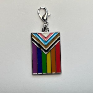 Pode incluir: Um amuleto de prata com um design de bandeira do arco-íris do orgulho. A bandeira tem um chevron preto apontando para baixo com um chevron marrom, azul, rosa e branco no topo. As listras do arco-íris são roxo, azul, verde, amarelo, laranja e vermelho.