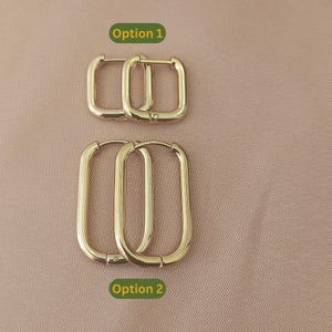 May include: Two pairs of gold-coloured hoop earrings. Option 1 features square hoops, while Option 2 showcases elongated oval hoops. The earrings are displayed on a neutral, pinkish-beige background.