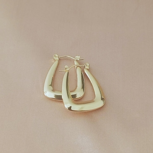 Op de afbeelding: Een paar goudkleurige, vierkante oorringen. De oorbellen hebben een gladde, gepolijste afwerking en een driehoekige vorm. De oorbellen worden gepresenteerd op een lichtroze oppervlak.