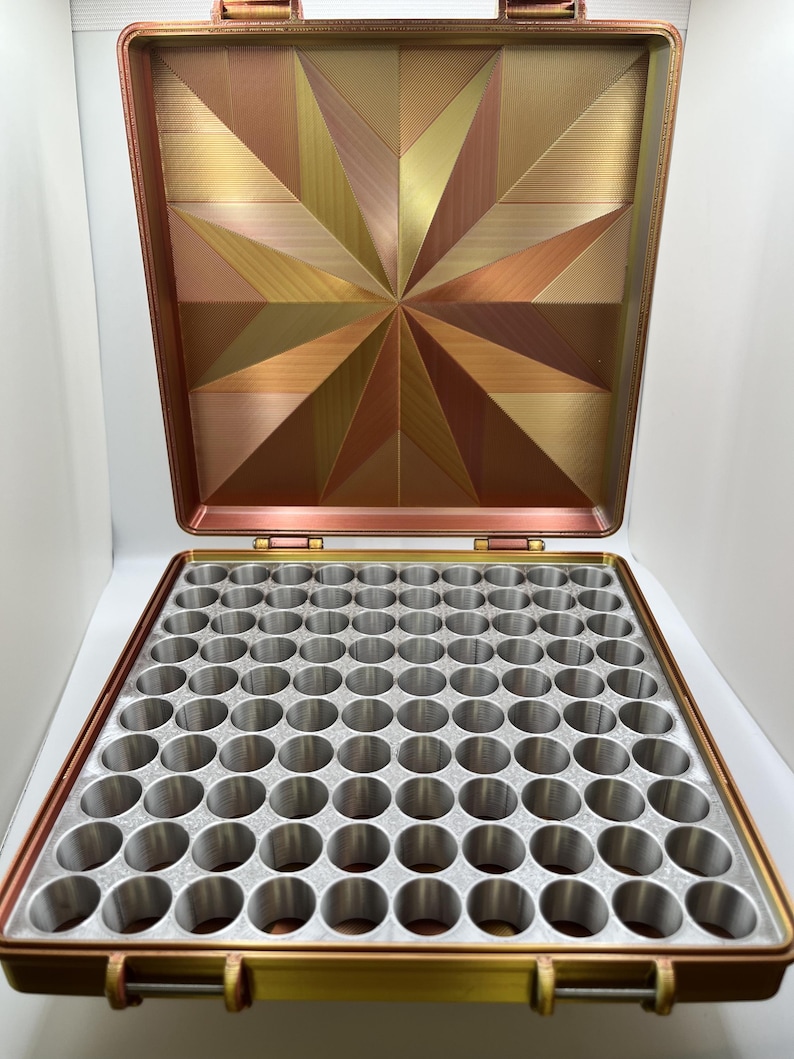 以下が含まれることがあります： 蓋に幾何学模様が施された、金色と銅色の収納ボックス。ボックスには、100個の丸い仕切りが付いた取り外し可能なトレイが付いています。