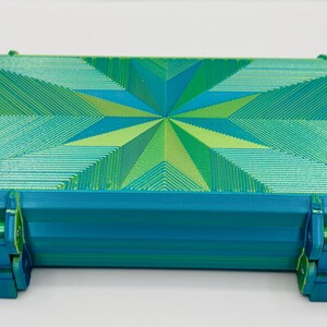 May include: A rectangular storage case featuring a vibrant geometric star pattern on the lid in shades of teal, lime green, and blue. The container has a textured surface with visible layered lines and two secure locking latches on the front edge.