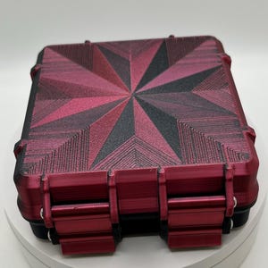 May include: A red and black rectangular storage case with a geometric pattern on the lid. The case has a latch on the front and a hinge on the back.