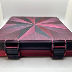 以下が含まれることがあります： 蓋に幾何学模様が施された赤と黒の収納ボックス。ボックスは両側に2つの留め具があります。