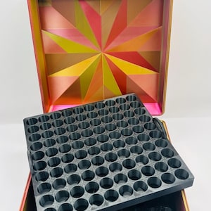 May include: A square storage case with a gradient pink, orange, and green exterior and a geometric starburst pattern on the interior lid. The case contains a removable black tray featuring a grid of circular compartments for small items.
