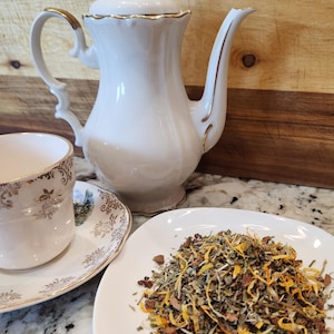 May include: A white teapot with gold trim and a white saucer with gold trim. A white teacup with gold trim and a floral design sits on the saucer. A white plate with a pile of loose leaf tea on it.