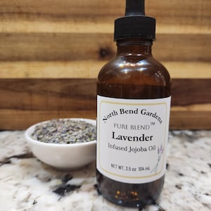 May include: A brown glass bottle with a dropper top, labeled "North Bend Gardens Pure Blend Lavender Infused Jojoba Oil" with a net weight of 3.5 oz or 104 ml. A small white bowl filled with dried lavender flowers is in the background.