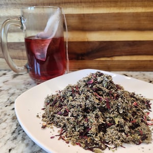 May include: A white plate with a pile of loose leaf tea blend. The tea blend is a mix of green, brown, and red herbs and flowers. A glass mug with a tea bag is in the background.