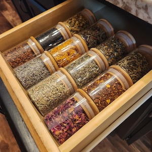 May include: A wooden drawer filled with glass jars containing various dried herbs and flowers. The jars are arranged in rows and have wooden lids.