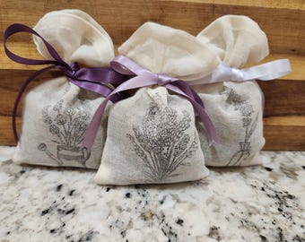 May include: Three small, off-white fabric sachets tied with purple and white ribbons. Each sachet is printed with a black and white illustration of lavender plants. The sachets are arranged on a speckled countertop.