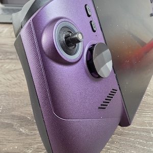 May include: A handheld gaming console featuring a metallic purple shell with a textured grip surface. The device includes a black joystick, a circular directional pad, two small oval buttons, a speaker grille, and a large rectangular screen.