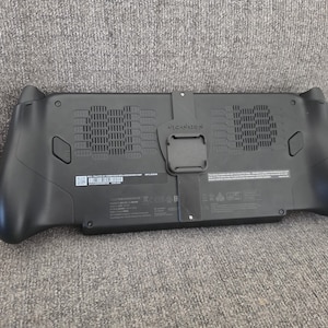 May include: A black handheld gaming console back panel featuring a central mounting bracket, two large ventilation grilles with horizontal slats, two rear-facing buttons, and various regulatory text markings printed on the lower center section.