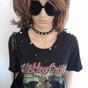 以下が含まれることがあります： 黒地にダメージ加工が施されたTシャツで、バンド「Motley Crue」のグラフィックプリントが施されています。首元にはシルバーのスタッズが施され、シルバーのスタッズが付いたブラックチョーカーネックレスが着用されています。