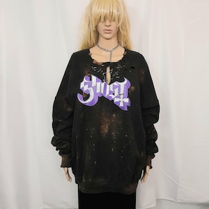 May include: Black distressed sweatshirt with a purple and white graphic that reads "Ghost". The sweatshirt has a crew neck with distressed detailing and long sleeves. The garment has a bleached, speckled design.