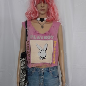 Puede incluir: Top sin mangas rosa con bordes desgastados y la palabra "PLAYBOY" impresa en la parte delantera. El top presenta un gráfico de un conejo con pajarita. Llevado con una falda vaquera y un bolso tote con estampado de cebra.